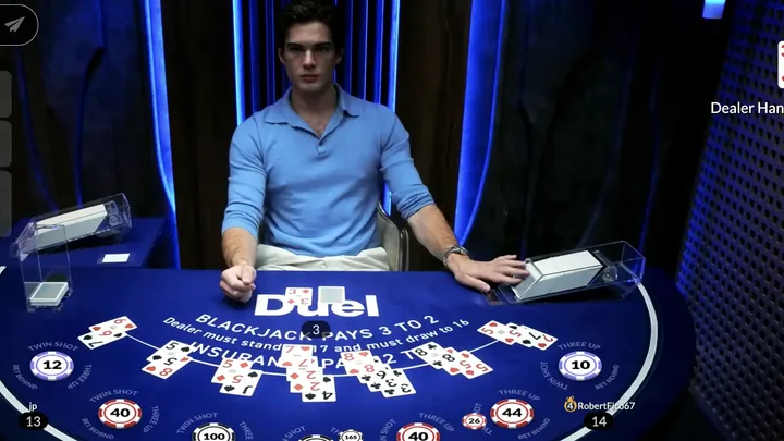 Dealer behind the shoe at the Duel Blackjack live table with seven boxes dealt across the blue felt